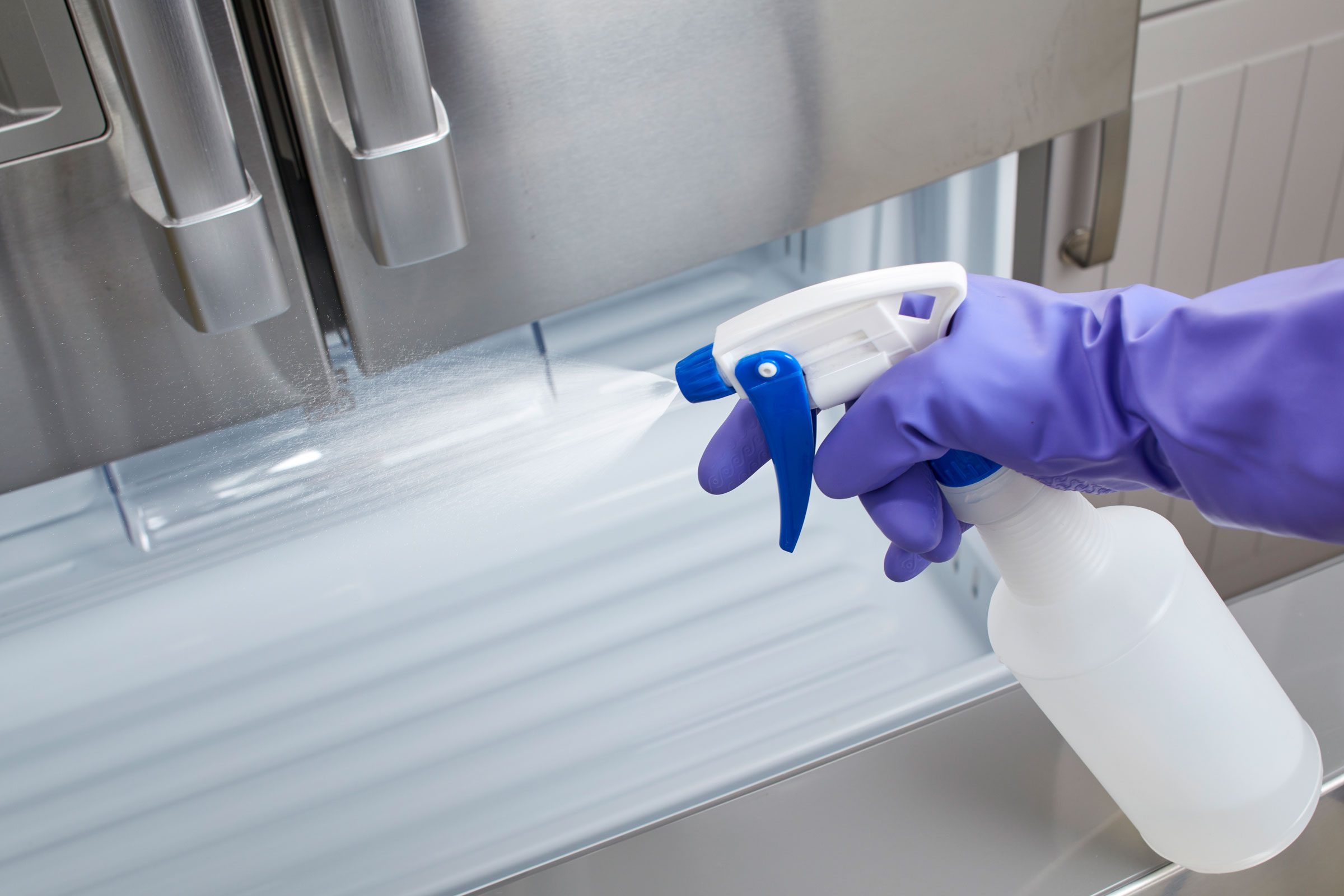gloved hand spraying cleaning fluid into open, empty freezer drawer