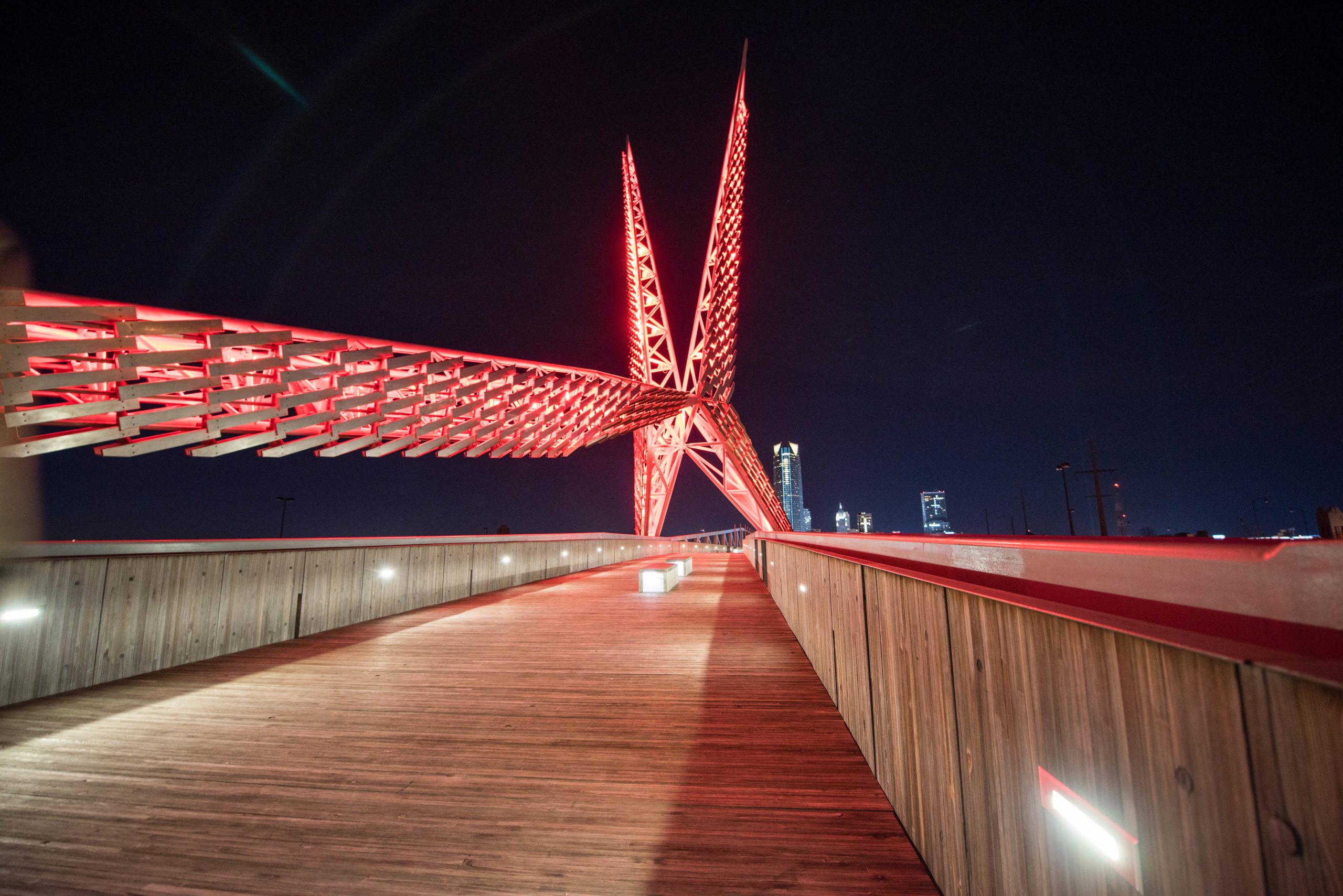 Bridge illuminated in red, extending into a night skyline. Wooden pathway and railing guide the view towards city buildings in the distance.
