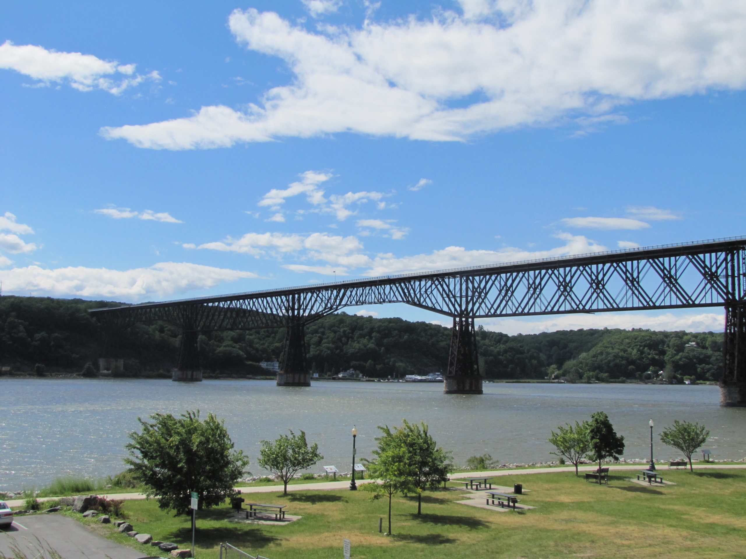 Bridge spans river, connecting tree-covered hills. Green park with picnic tables and cars in foreground, beneath bright blue sky with scattered clouds.