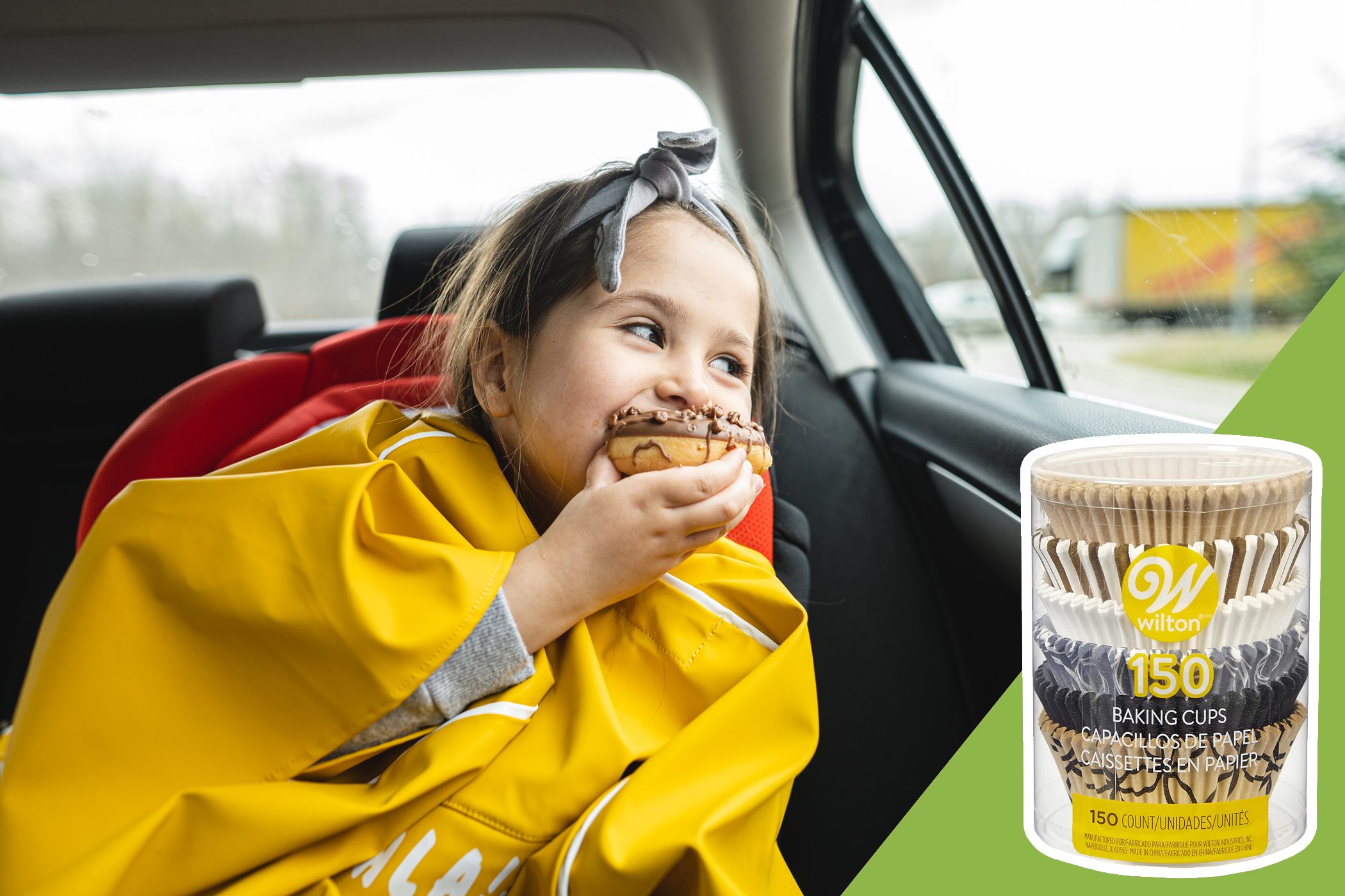 A child enjoys a donut in the backseat of a car, wearing a yellow raincoat. Nearby, there's a package labeled "Wilton 150 Baking Cups."