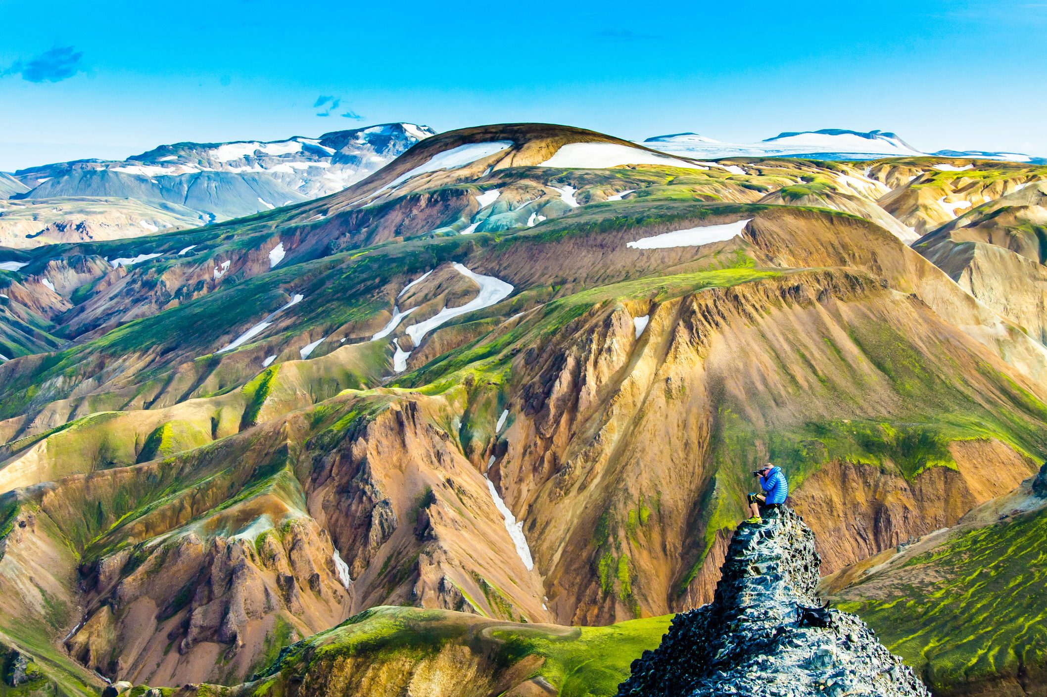 Person sitting, taking a photograph, on a rocky outcrop in a colorful, mountainous landscape with patches of snow and a clear blue sky.