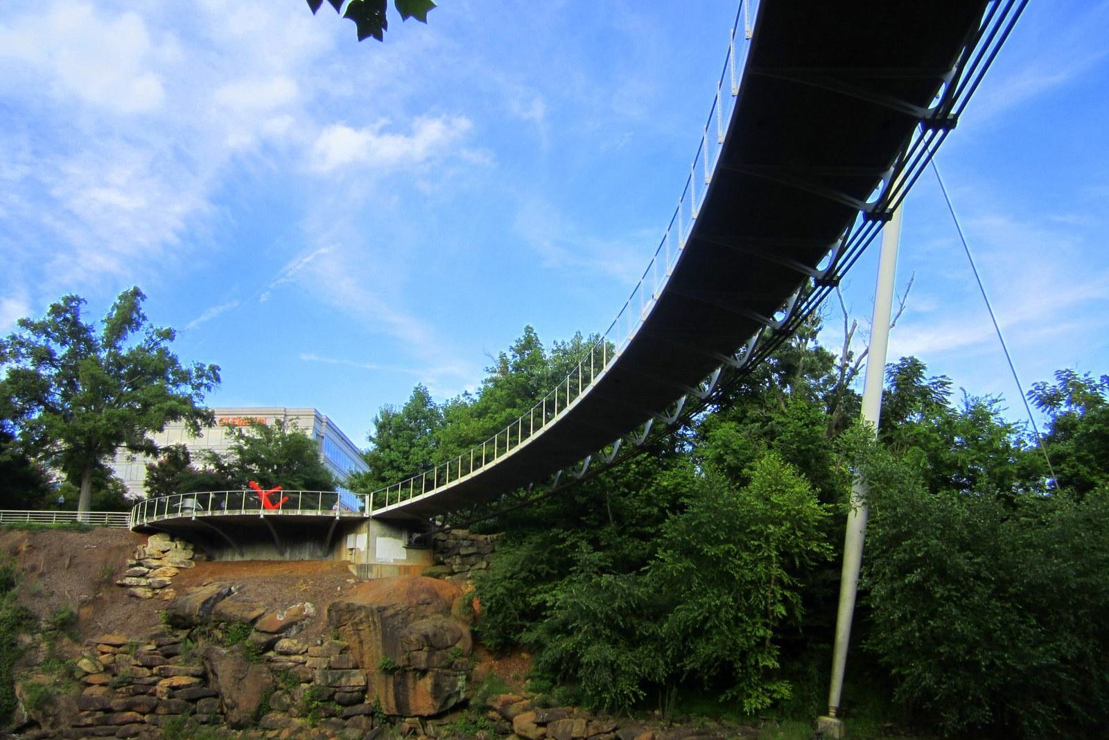 Curved pedestrian bridge spans over lush green trees and rocky terrain, under a bright blue sky.