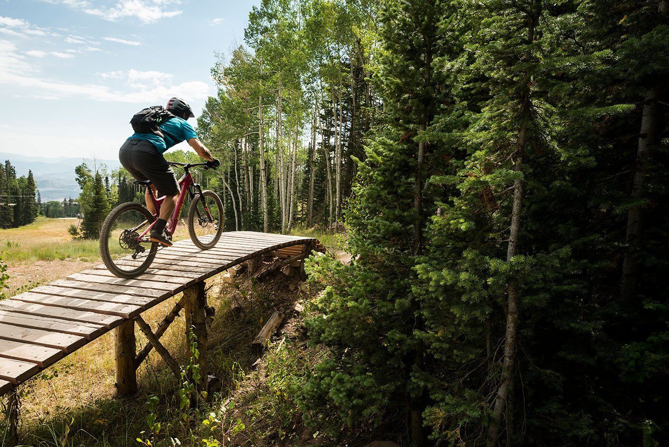 Cyclist rides wooden bridge through forested trail with tall trees under a clear sky.