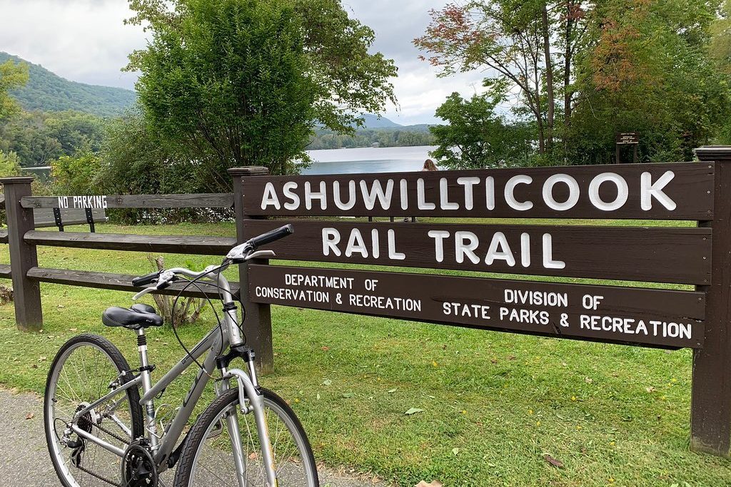 Bicycle rests against a wooden sign reading "Ashuwillticook Rail Trail," amidst lush greenery and a distant mountainous landscape.