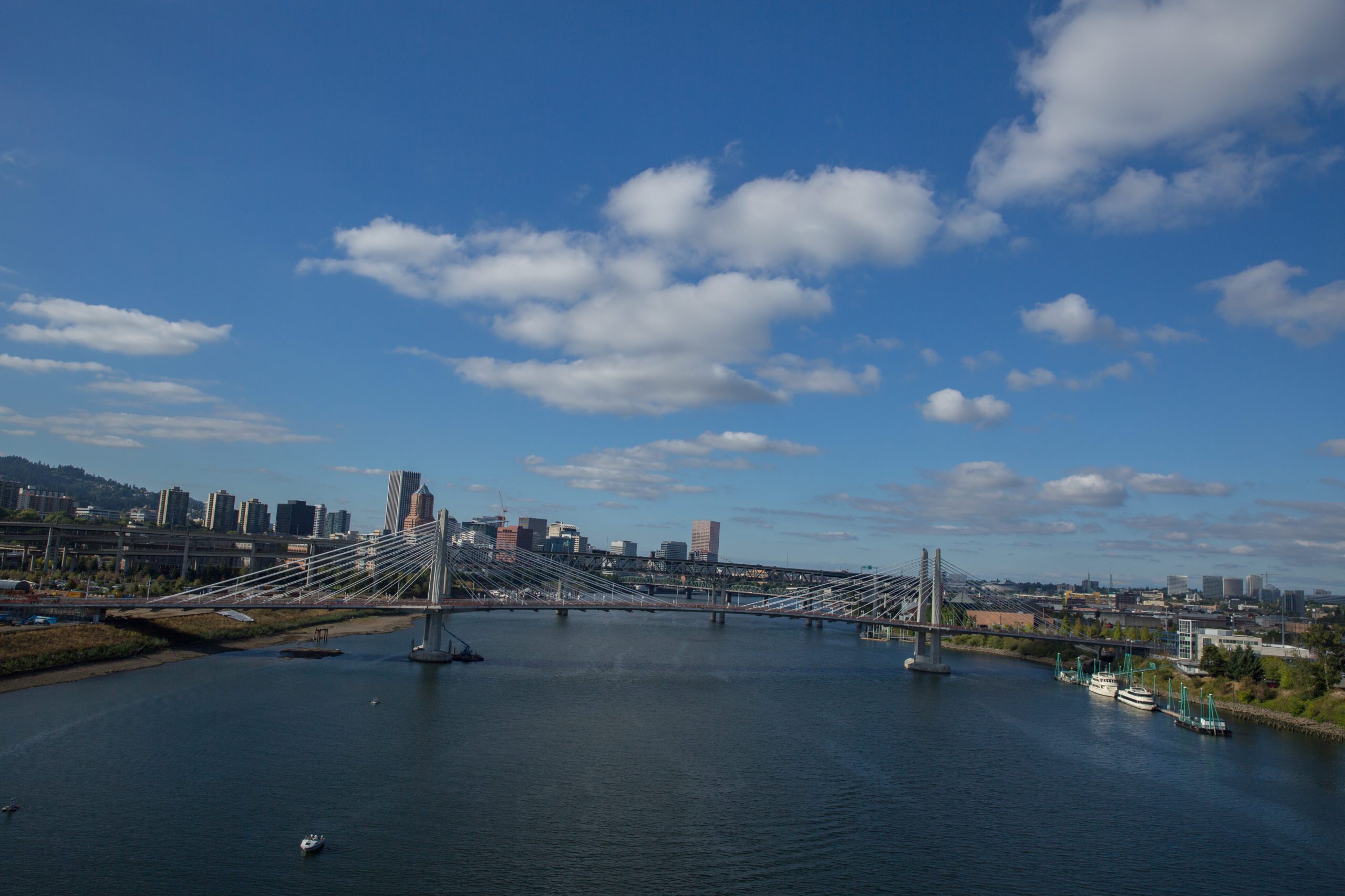 Bridge spans a river beneath a partly cloudy sky, with a city skyline in the background and boats visible on the water.