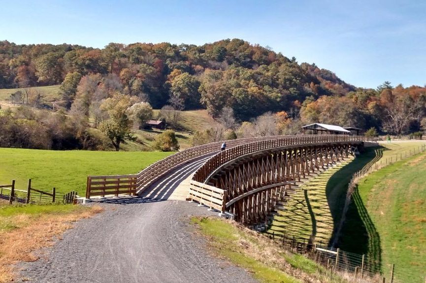 A cyclist crosses a wooden bridge on a path through rolling green fields with a wooded hillside in the background.