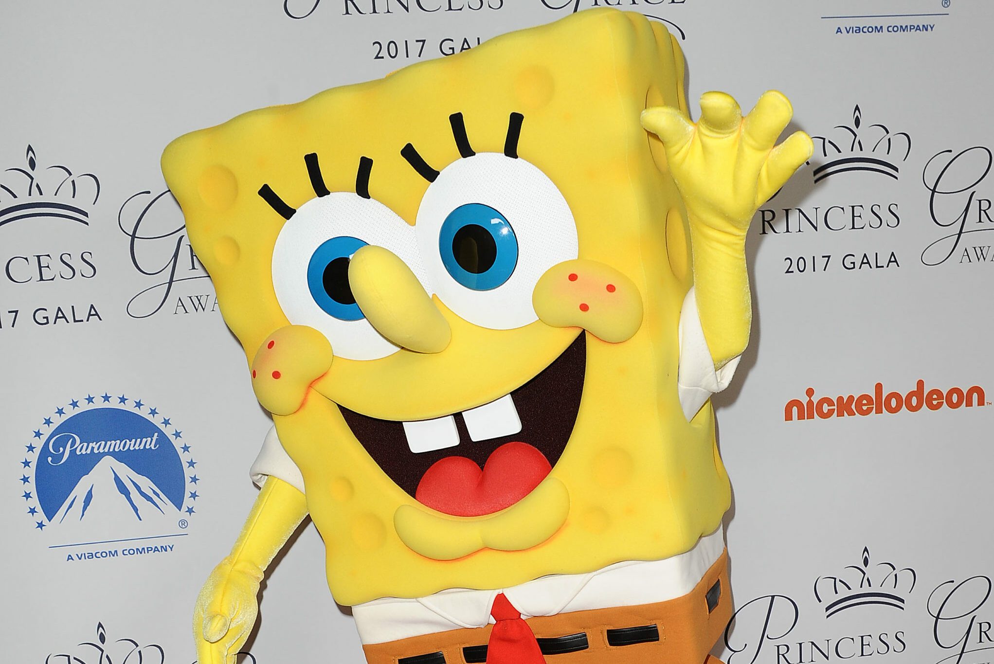 A large yellow sponge character waves energetically at a formal event, with logos and text on the backdrop indicating a gala and media companies.