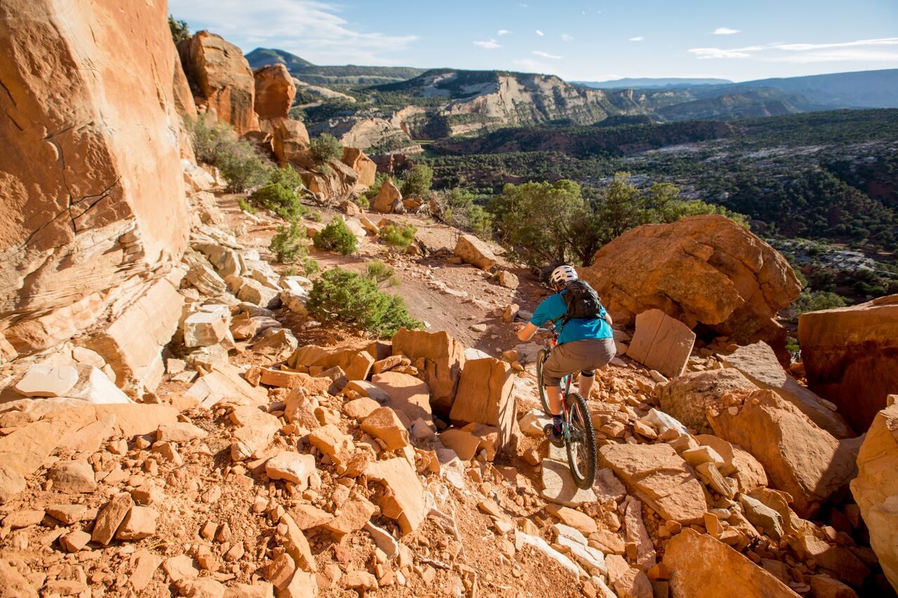 Cyclist navigating rocky trail on a mountain bike, surrounded by large reddish boulders and cliffs under a clear blue sky.