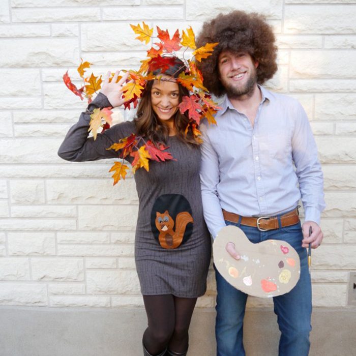 Two people pose; one wears a dress with a squirrel and leaf headpiece, the other holds a paint palette, standing against a brick wall.