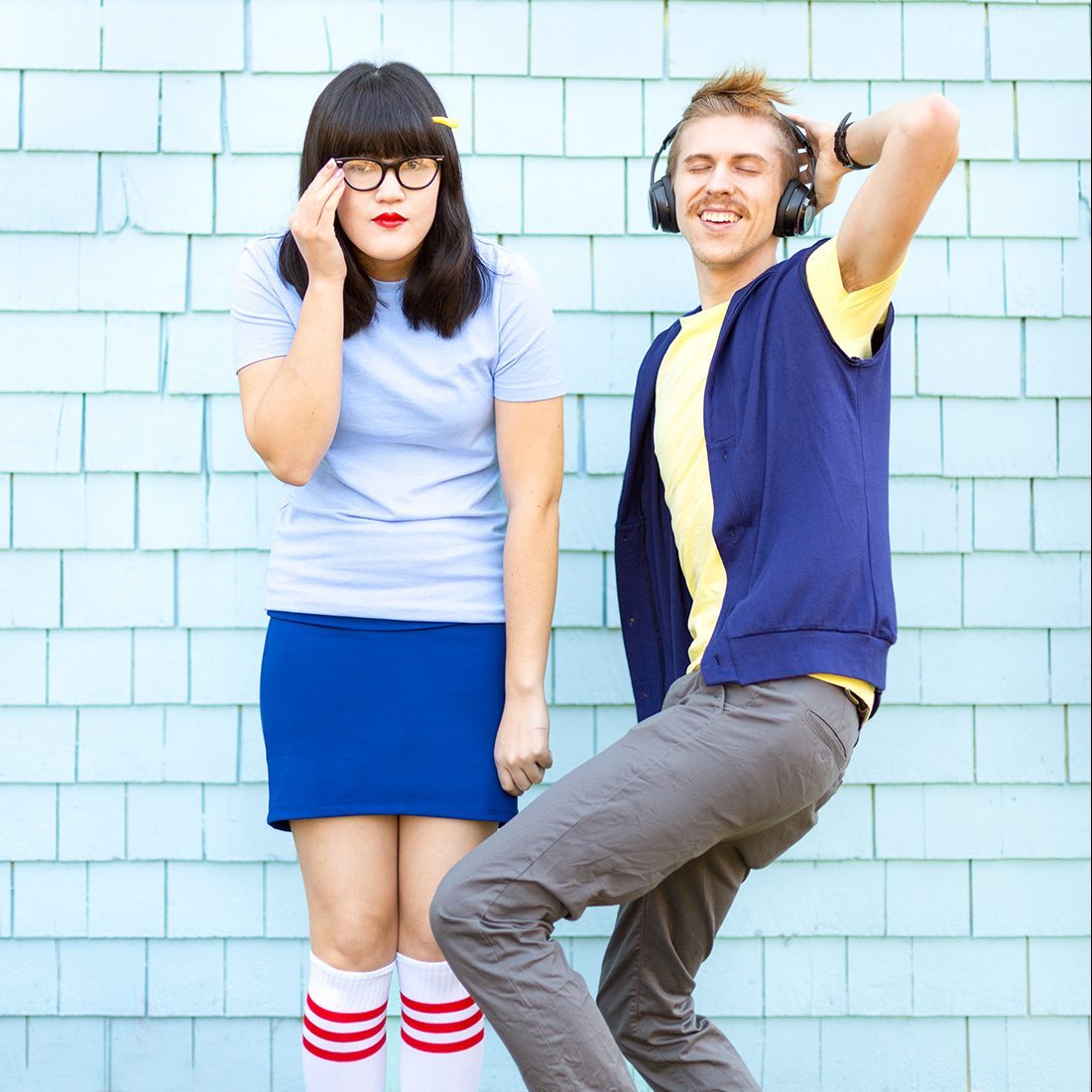 Person adjusting glasses stands beside person dancing with headphones against light blue shingled wall.