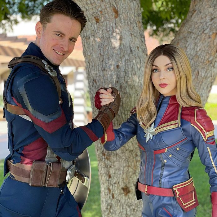 Two people in superhero costumes shake hands, standing in front of a large tree in a sunny outdoor setting.