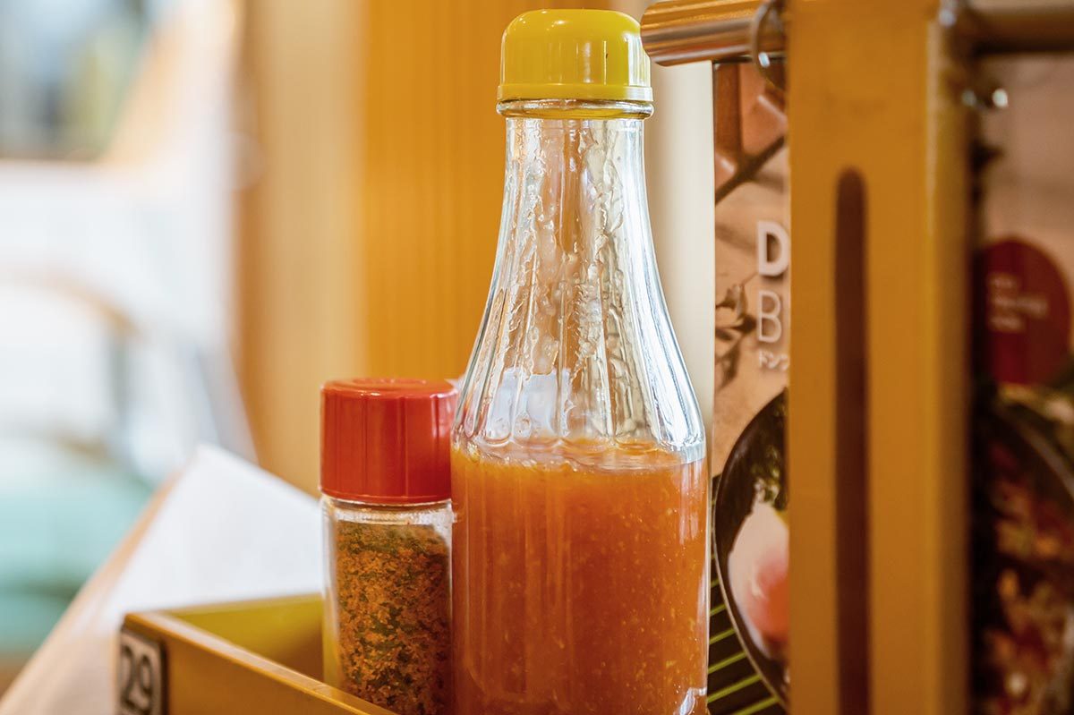 Glass bottle holds orange sauce, standing beside small spice jar, inside a wooden condiment holder on a restaurant table.
