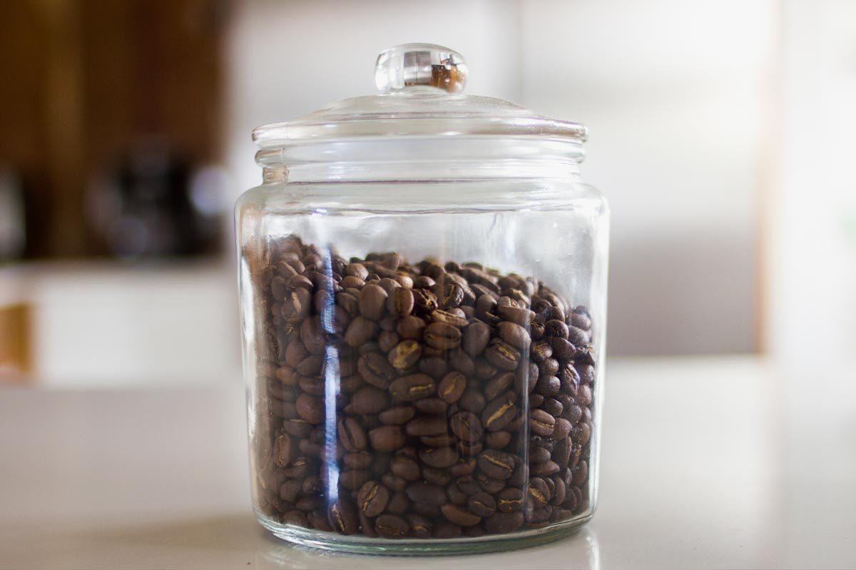 Glass jar holds coffee beans, sitting on a white countertop in a softly lit kitchen environment.