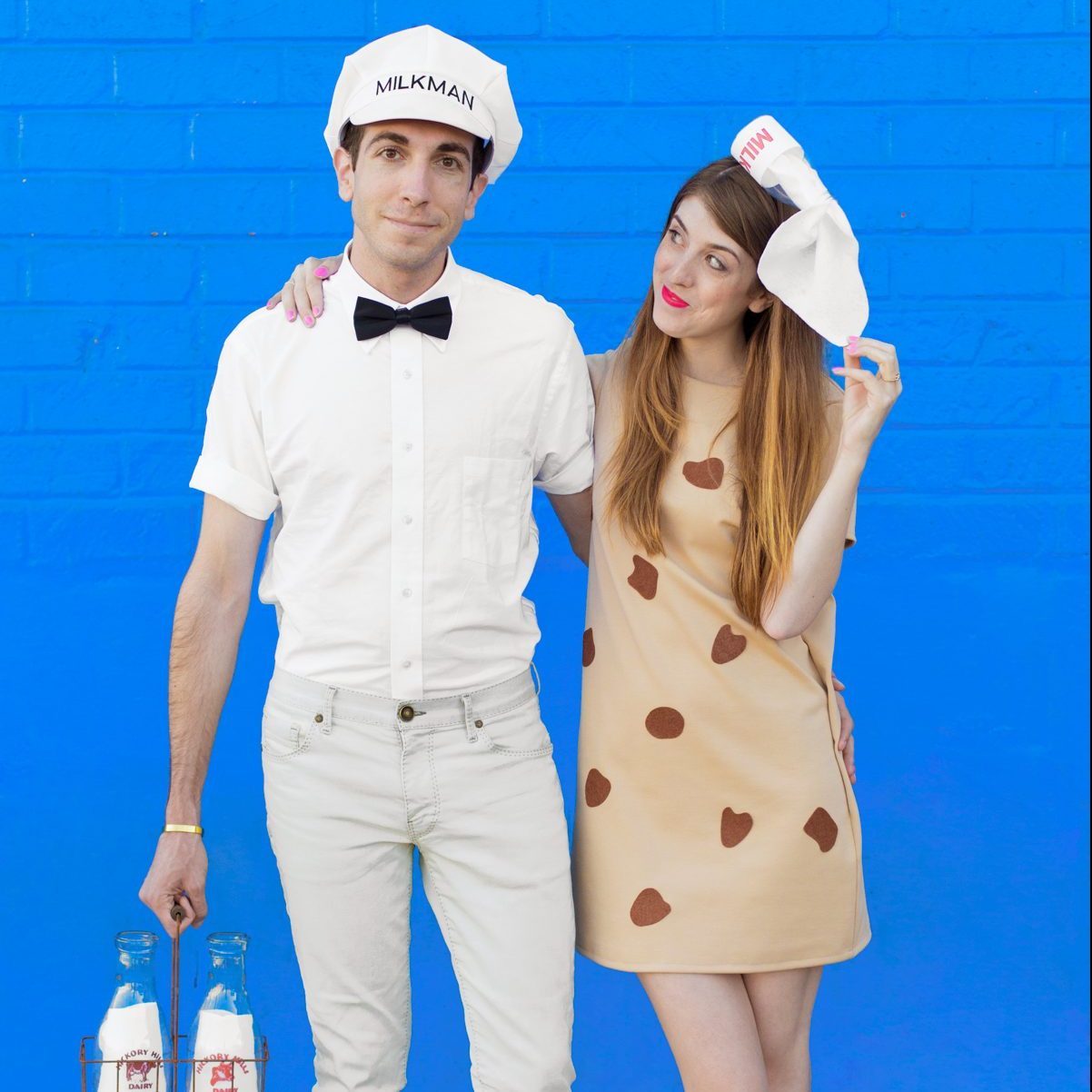 Man dressed as a milkman and woman as a cookie, posing against a bright blue wall. He holds a milk bottle carrier.