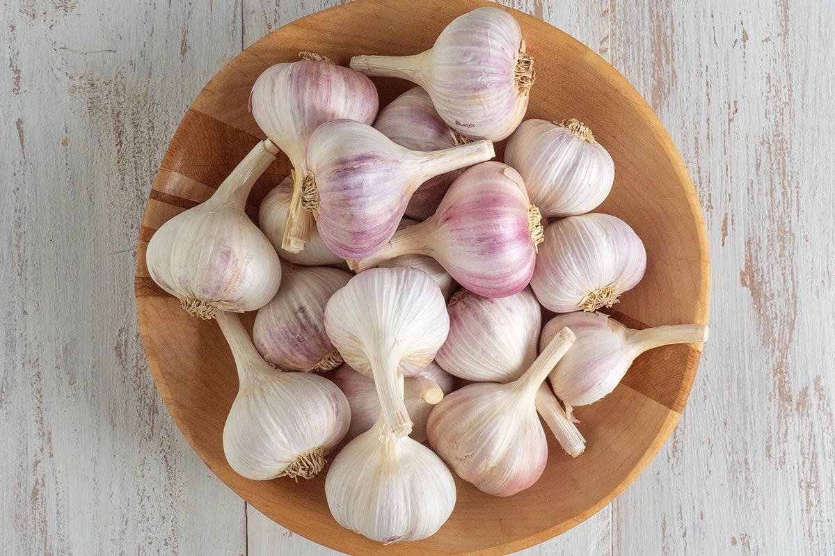 Garlic bulbs rest in a wooden bowl on a rustic, light-colored wooden surface.