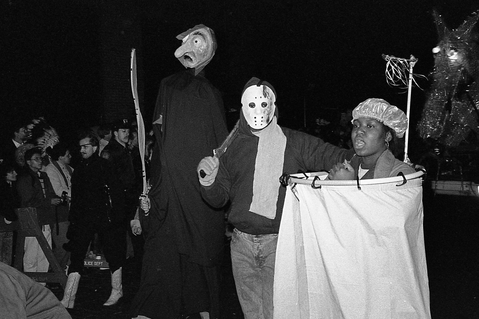 A crowd celebrates a costume parade at night; figures in masks and robes, a person in a shower costume, spectators with diverse outfits.