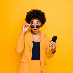 Person holding sunglasses and phone, expressing surprise, against a vibrant yellow background.