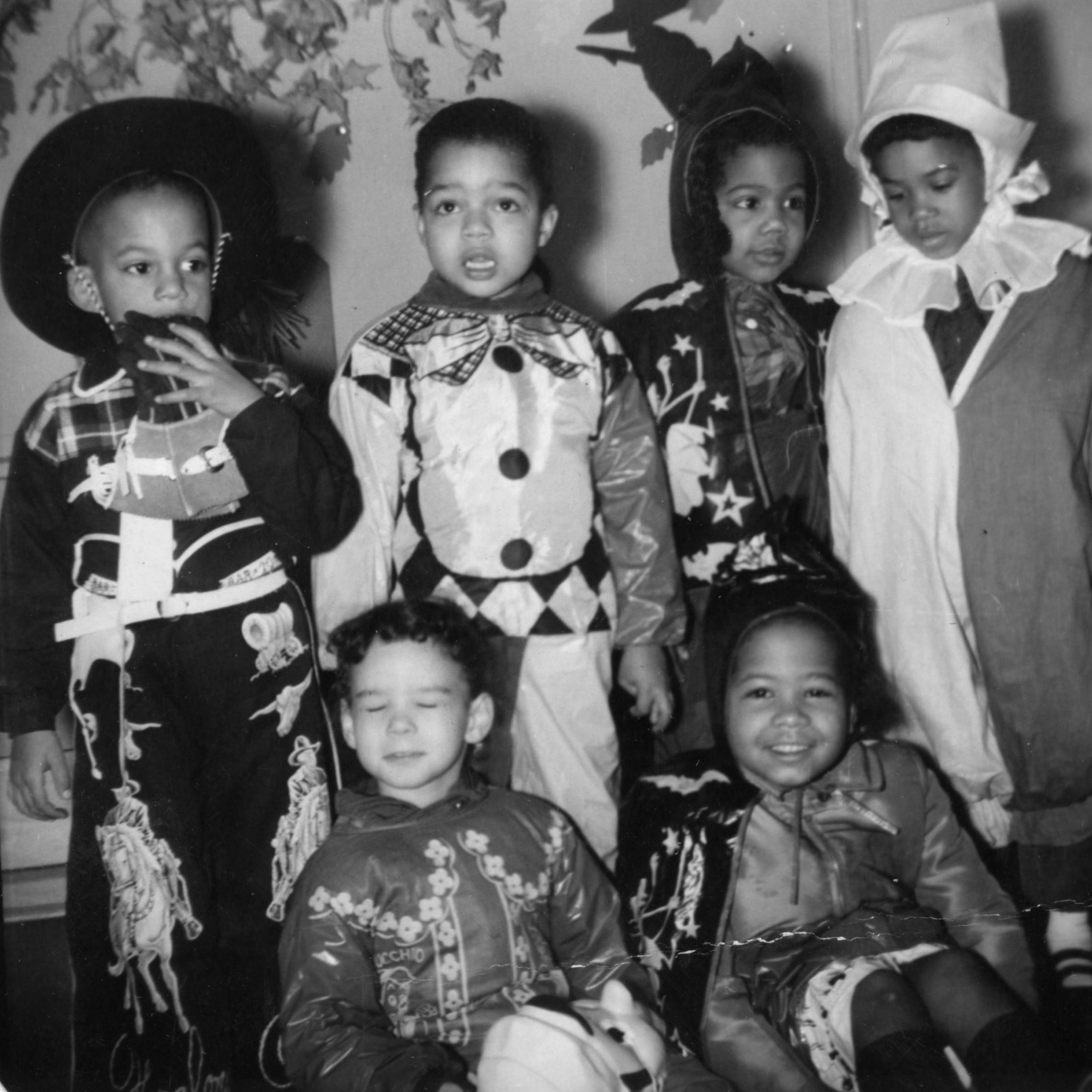Children dressed in costumes stand and sit together, smiling. They appear to be at a festive event, with decorative leaf patterns on the wall behind them.