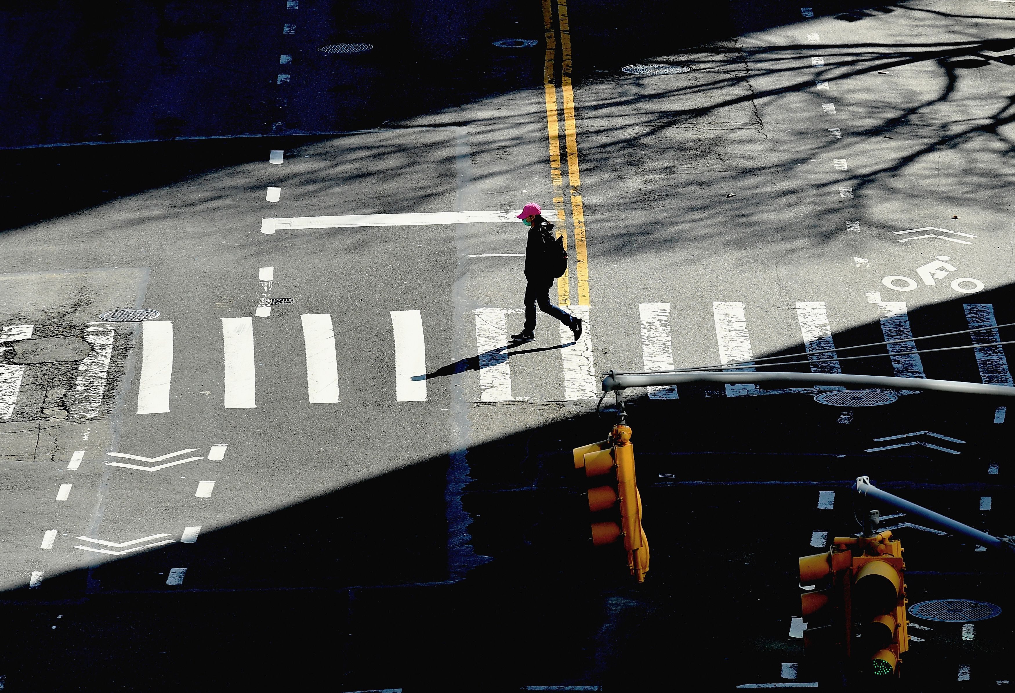 A person walks across a sunlit crosswalk on an empty city street, casting a shadow, with traffic lights and road markings visible.