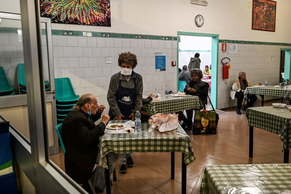 People eat at tables in a cafeteria setting; one person stands wearing a "SANT'EGIDIO" apron. Green checkered tablecloths, white-tiled walls surround.