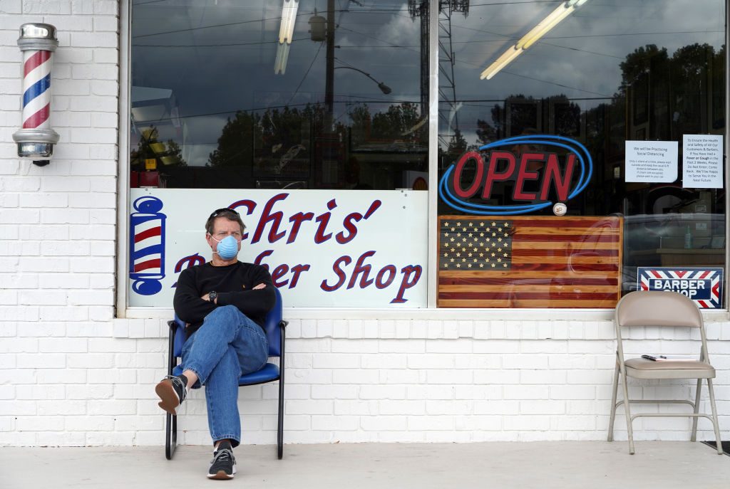 Man wearing mask sits on chair outside "Chris' Barber Shop" with an open sign and American flag in window. White brick exterior with barber pole.