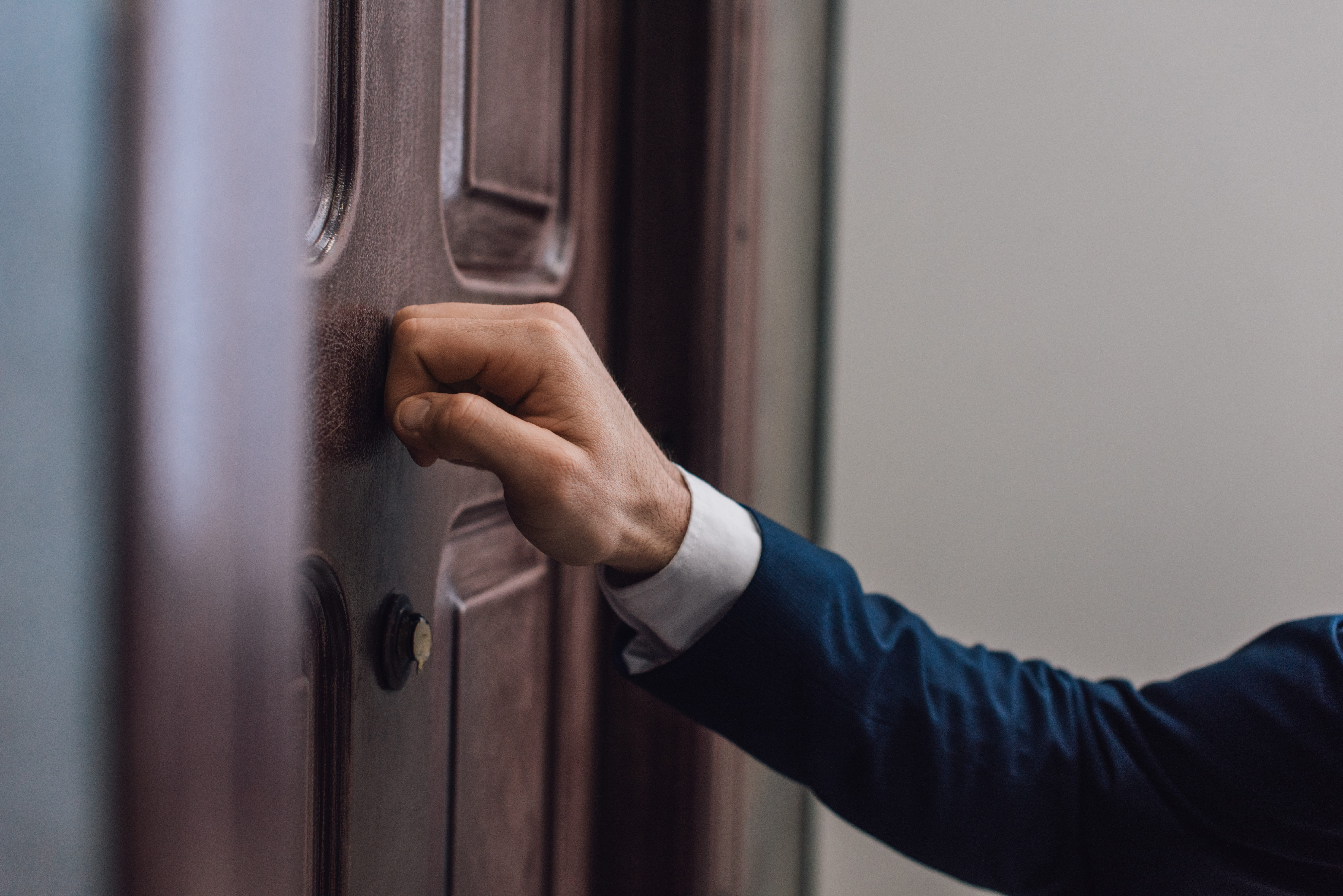 A hand knocks on a wooden door in a hallway, wearing a dark suit jacket.