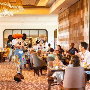 Mickey Mouse waves in a vibrant, modern restaurant as families dine and applaud, with another character approaching in the background.