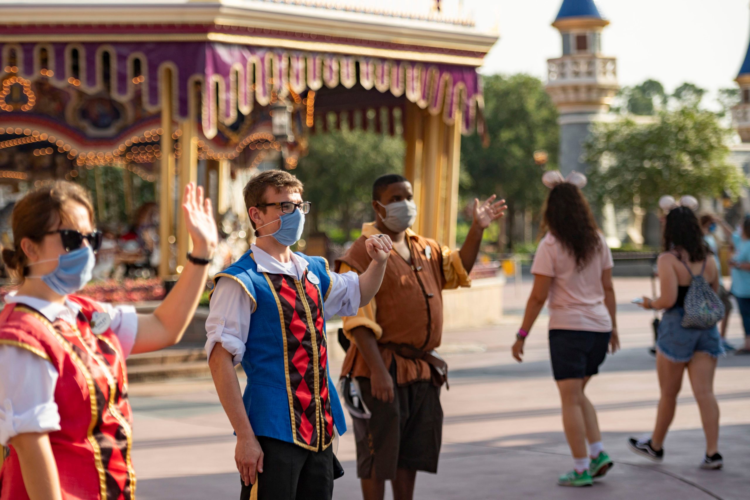 People wearing costumes and masks welcoming visitors in a theme park, near a carousel with decorative lights and towers in the background.
