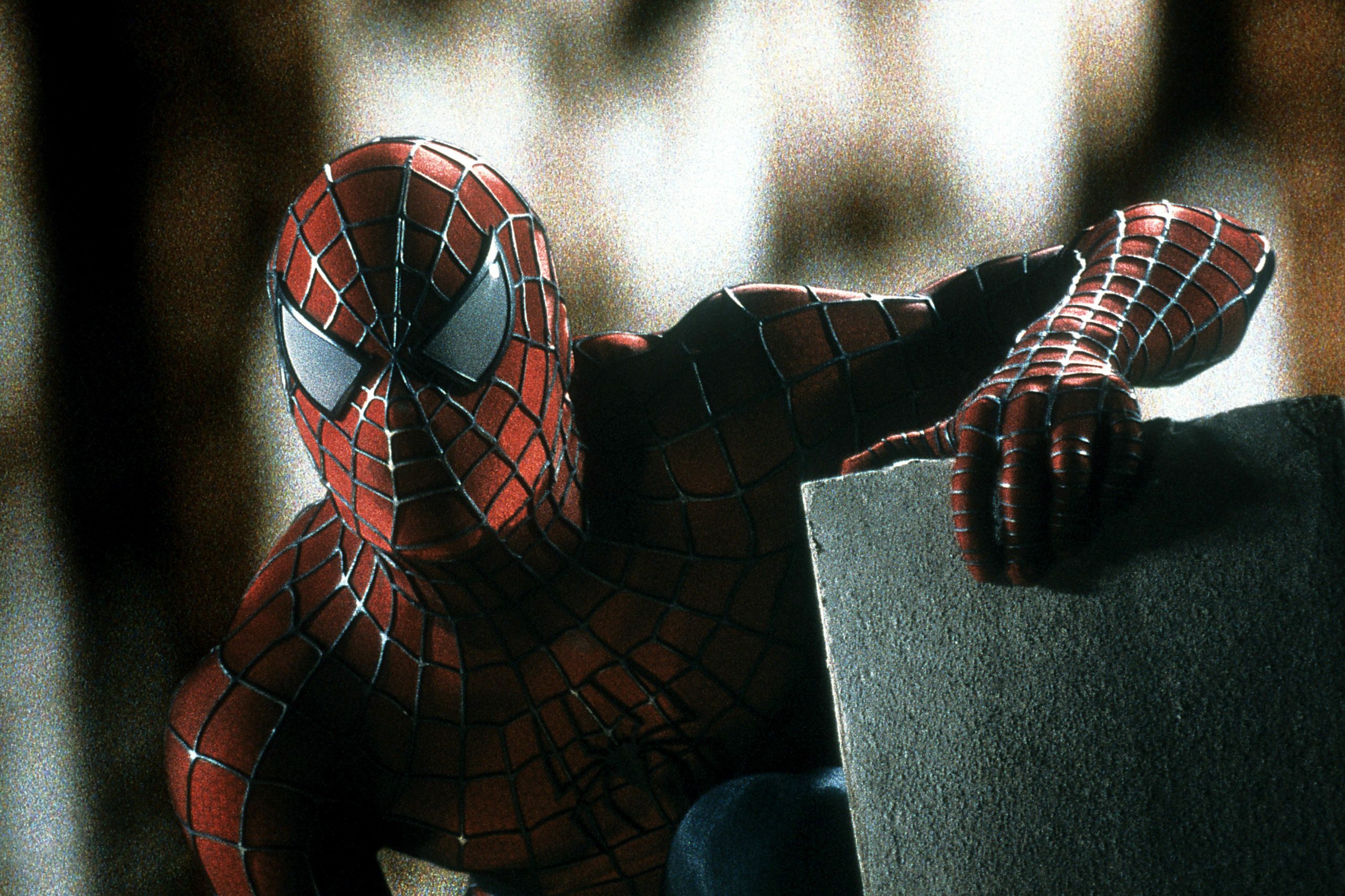 A figure in a red and blue suit crouches, gripping a stone ledge, against a blurred urban background.