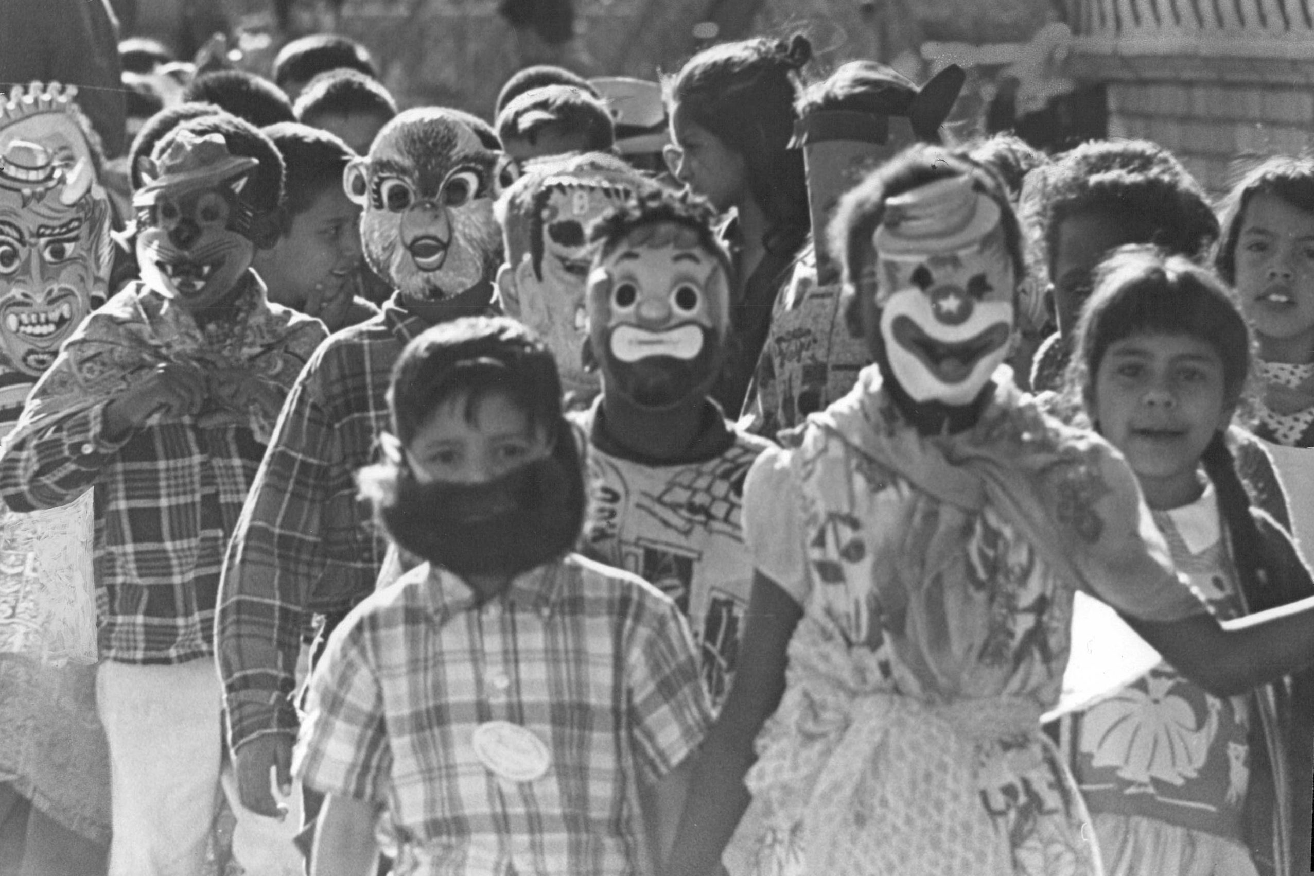 Children wear various masks, walking closely together, in a festive outdoor gathering.