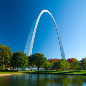 Arch reflecting in water, surrounded by trees and green grass, with a clear blue sky.