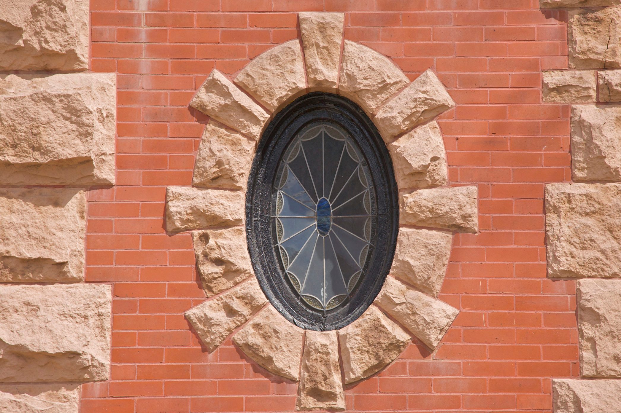 Oval stained glass window surrounded by rough stone blocks, set in a red brick wall.