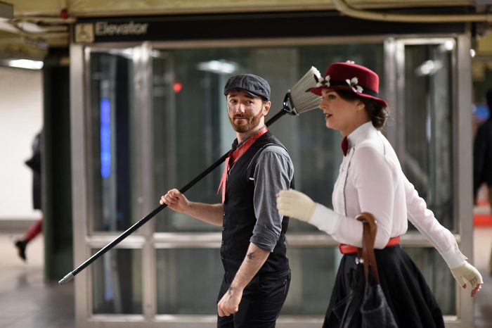 Man holds broom on shoulder, walking with woman in vintage attire, in a subway station beneath an 