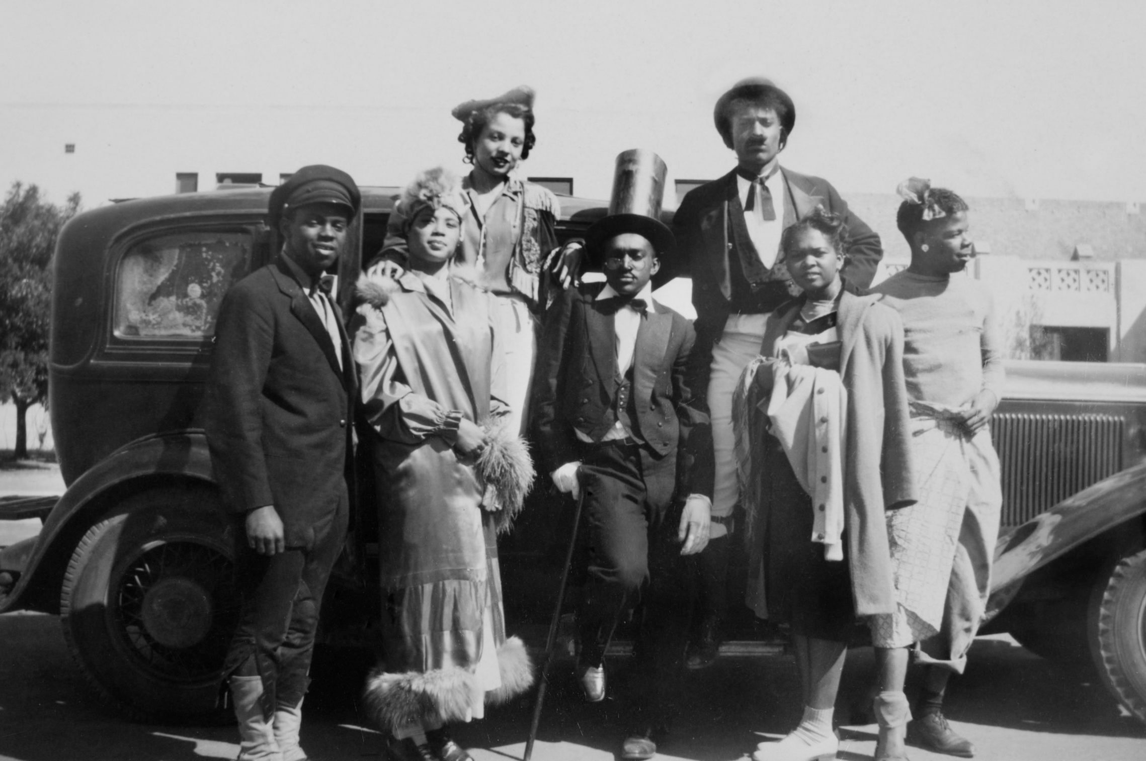 Seven people pose in vintage attire beside an old car outdoors, exuding a lively and stylish atmosphere under the midday sun.