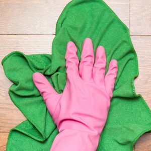 A pink-gloved hand wipes a green cloth on a wooden surface.