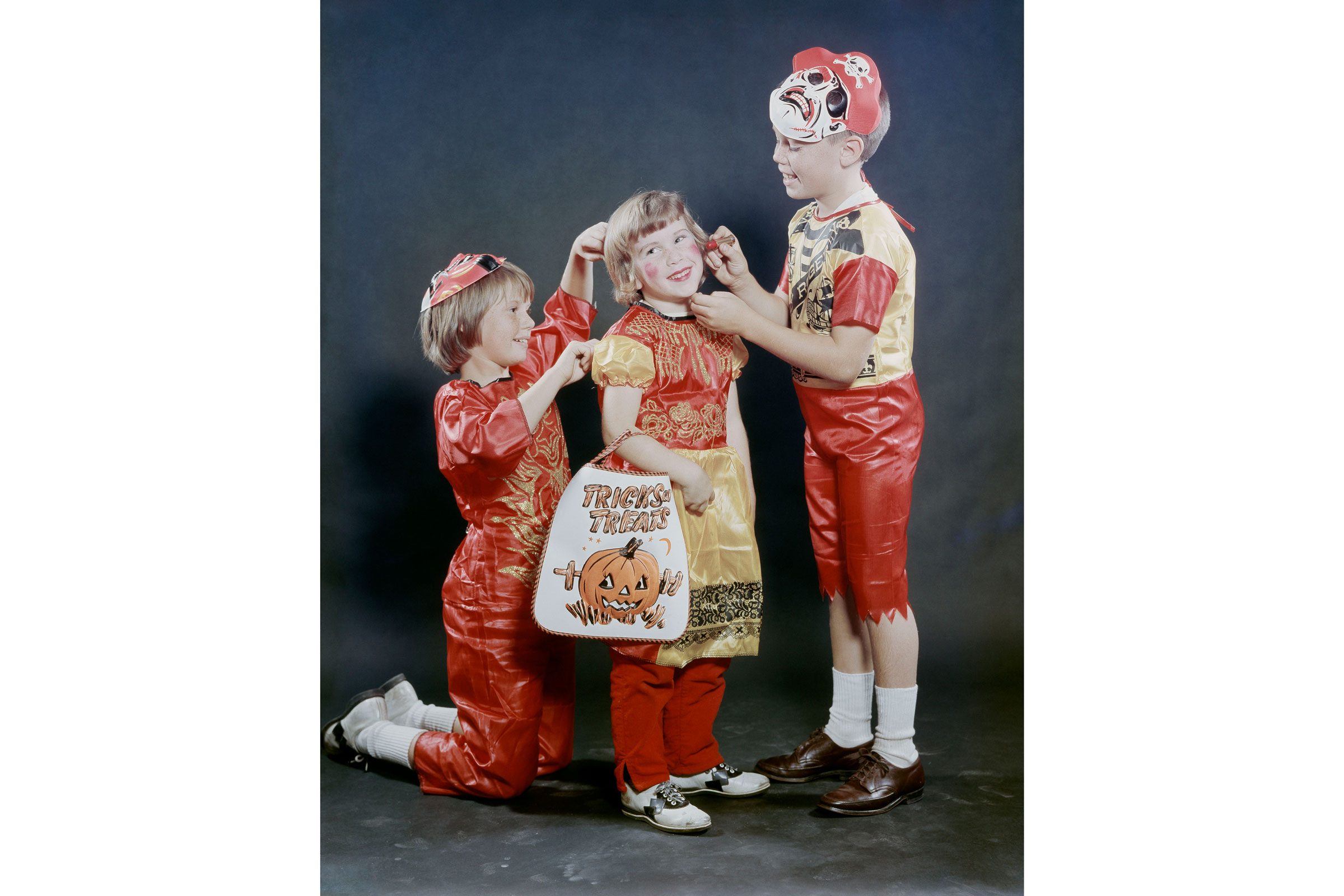 Three children in Halloween costumes adjust each other's accessories, holding a "Tricks or Treats" pumpkin bag, against a plain studio backdrop.
