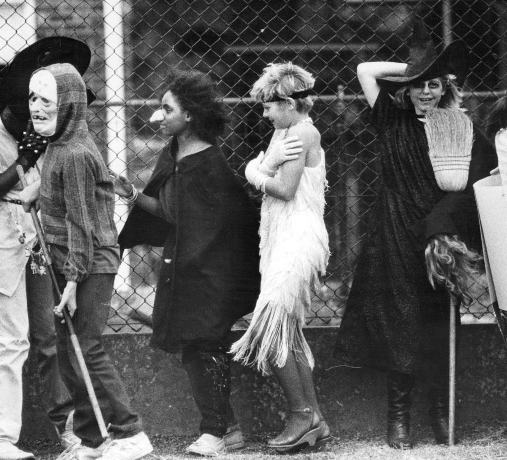 Children wear Halloween costumes, standing in line outdoors, beside a chain-link fence, showcasing various outfits like witches and monsters.