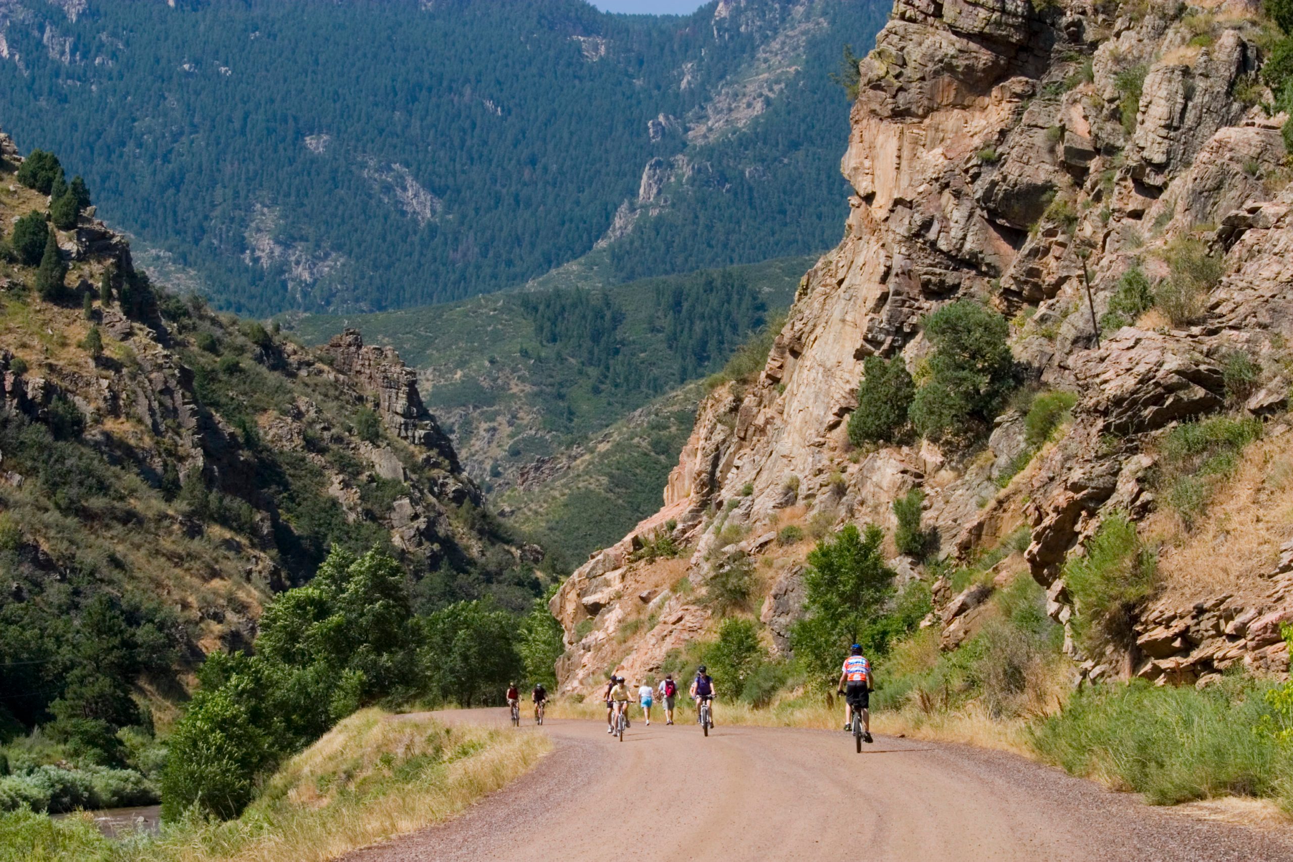 Cyclists ride on a winding dirt path through a rocky, mountainous landscape surrounded by green trees and steep cliffs.