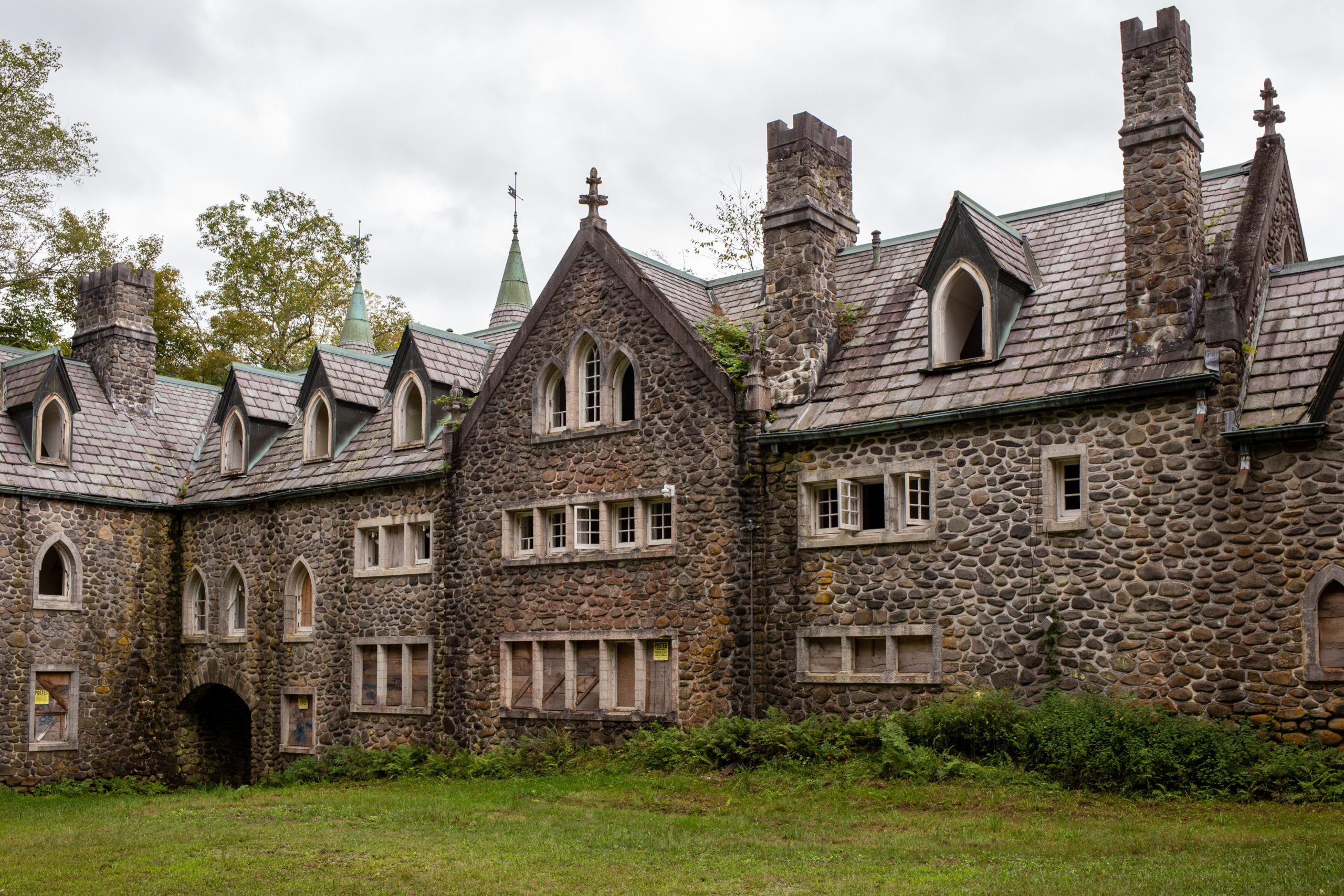 Stone building with arched windows stands surrounded by greenery and trees, under a cloudy sky.