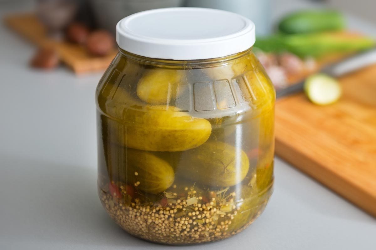 A jar holds pickles submerged in brine on a kitchen counter, with mustard seeds visible, near a cutting board and fresh cucumbers.