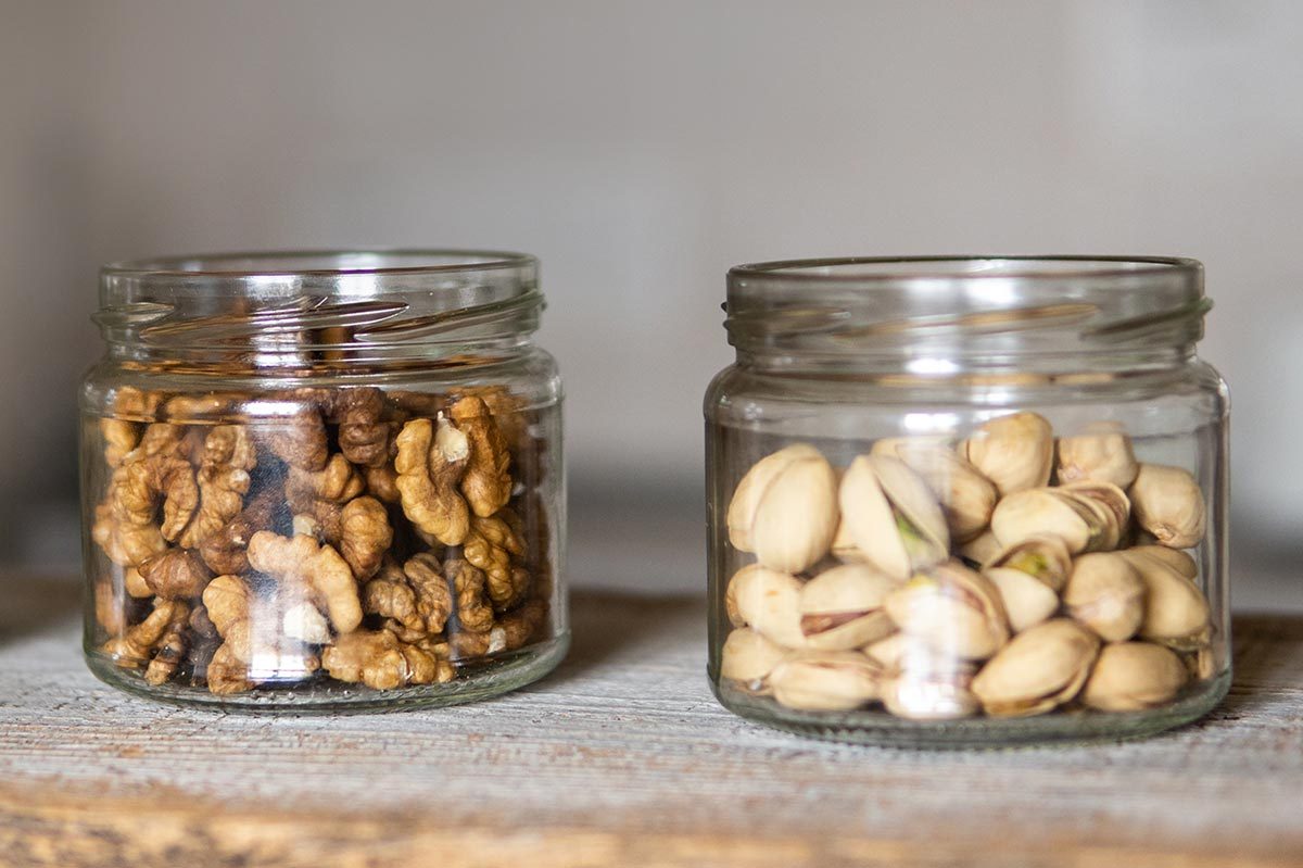 Jars hold walnuts and pistachios, sitting side by side on a wooden surface in a softly lit room.