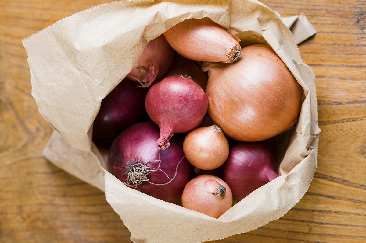 A variety of onions rest inside a brown paper bag on a wooden table.