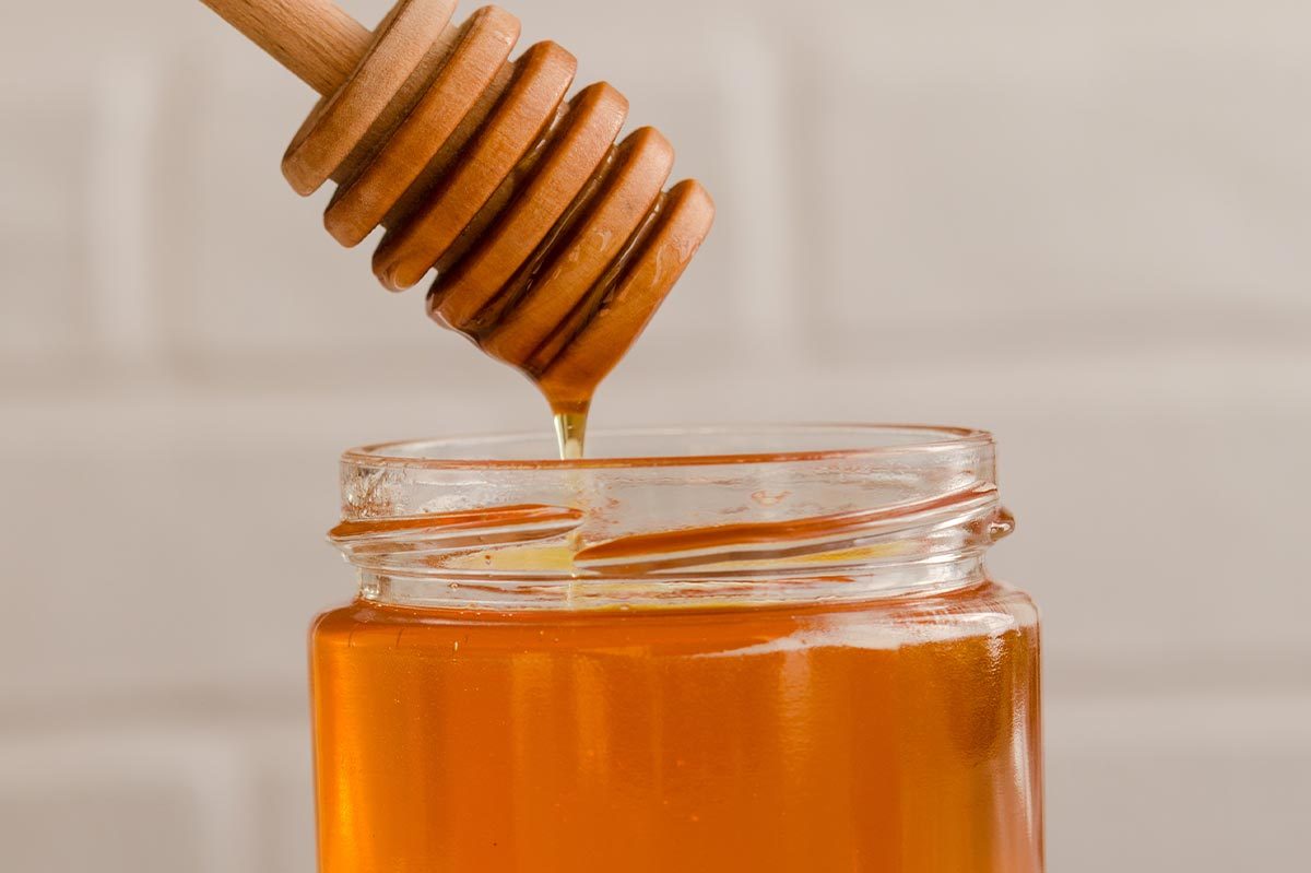 Honey drips from a wooden dipper into a glass jar, against a neutral background.