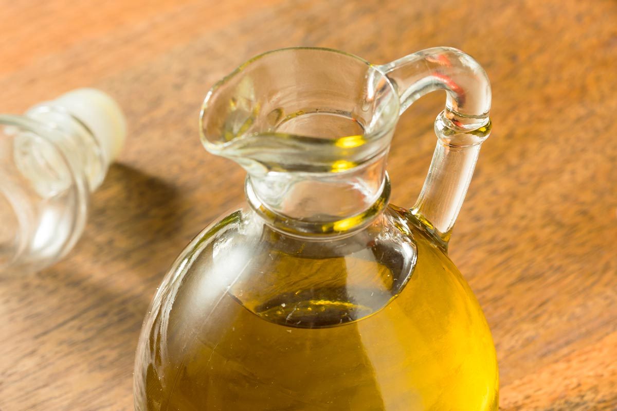Glass pitcher contains golden liquid, likely oil, on wooden table. Nearby, a bottle lies on its side, partially out of view.