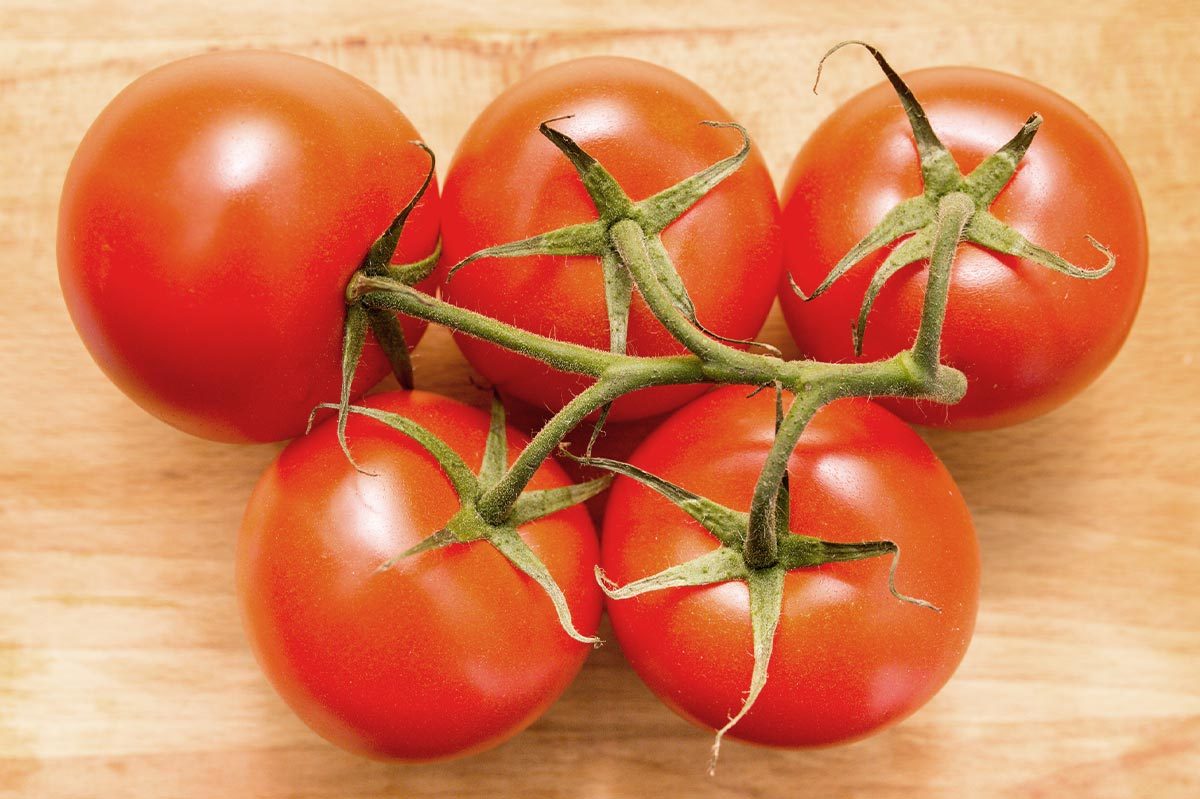 Five ripe tomatoes cluster together on connected green stems, resting on a light wooden surface.
