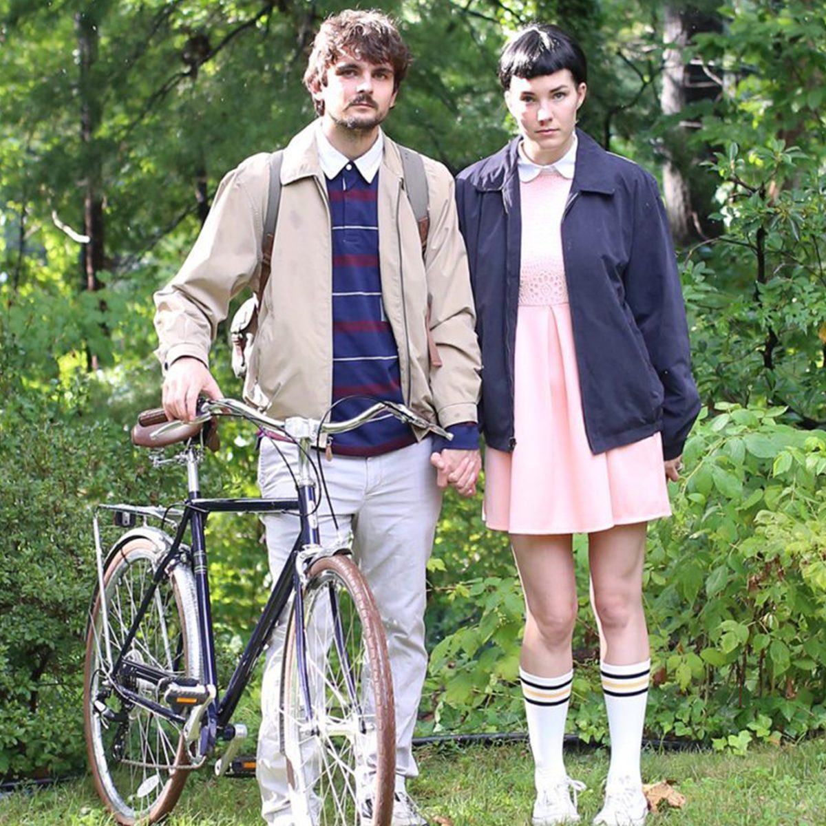 Two people stand holding hands beside a bicycle in a lush, green park.