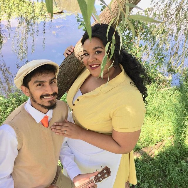 Two people are posing outdoors; one holds a ukulele and wears a cap, while the other leans against a tree near a pond.