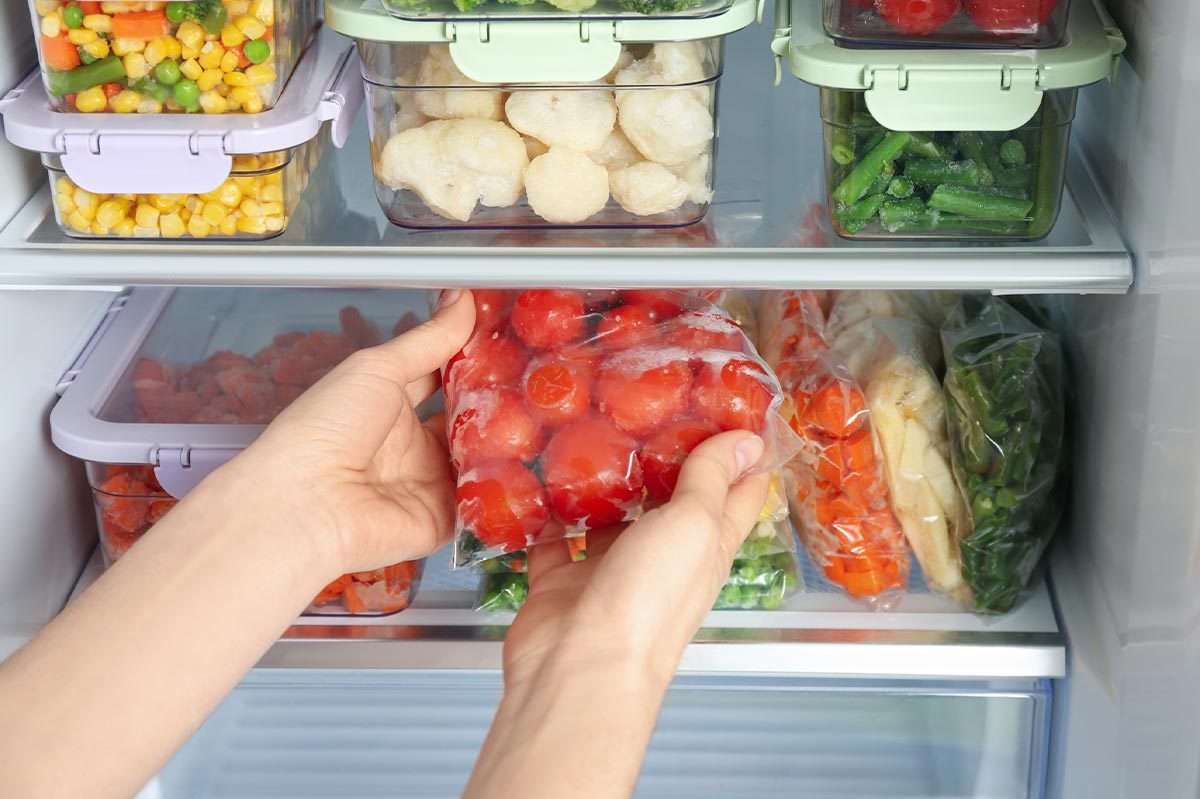 Hands holding bag of frozen cherry tomatoes in a freezer filled with various other frozen vegetables in containers and bags.