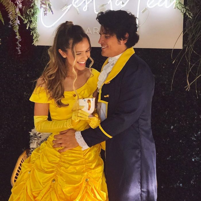 Couple dressed as Beauty and Beast smiling, holding Chip teacup, surrounded by greenery and illuminated sign.