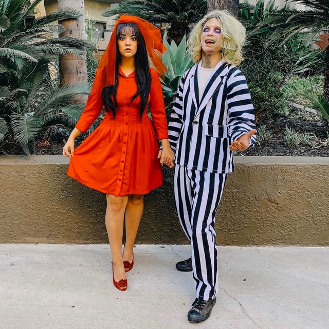 Two people in themed costumes stand together, holding hands, in an outdoor setting with plants and trees.